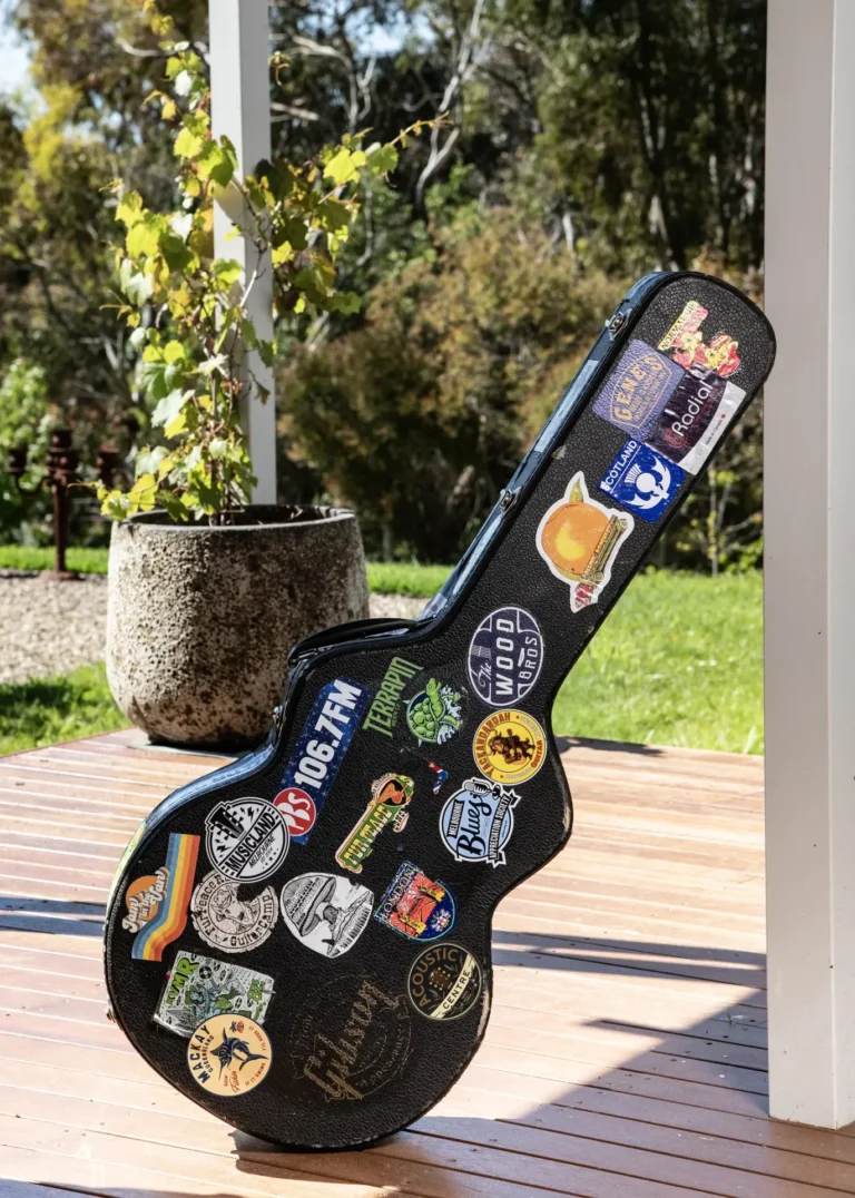 Sticker-covered acoustic guitar case resting on a timber deck in a quiet rural setting